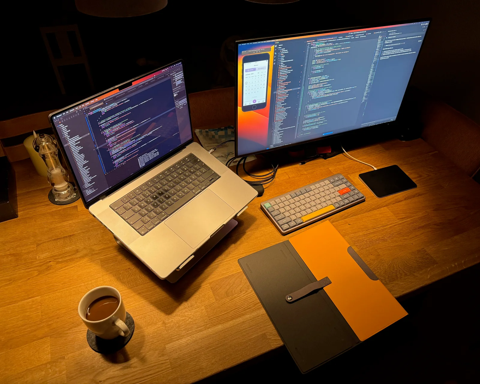 A softly lit developer workstation with a laptop, monitor, notebook and coffee